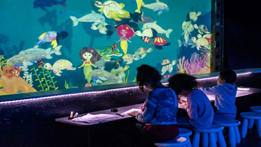 인어, 물고기, 바다 생물의 그림으로 덮인 바다를 테마로 한 벽 앞에 놓인 테이블에서 두 어린이가 색칠하고 있습니다.