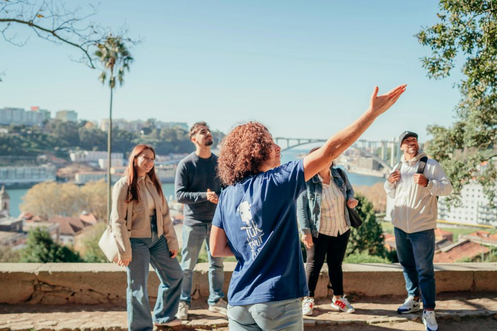 한 여성이 다리와 도시 풍경을 배경으로 야외에서 다섯 명의 사람들과 대화를 나누며 몸짓을 하고 있습니다.