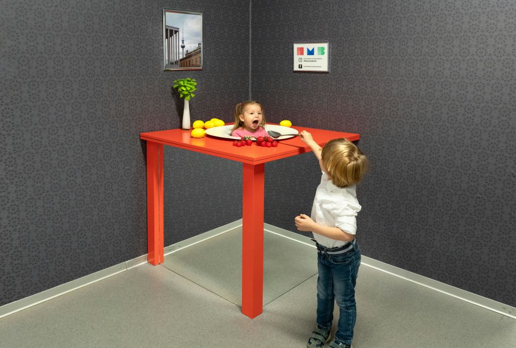 한 어린이가 장난스럽게 다른 어린이의 머리를 향해 손을 뻗고 있는데, 트릭 아트 전시품에 과일이 놓인 탁자 위에 놓여 있는 것처럼 보입니다.