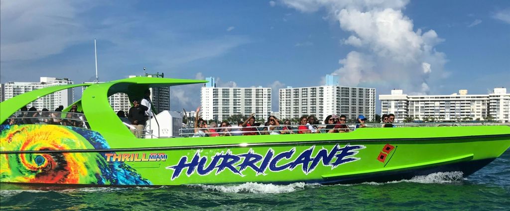 "허리케인" 이라는 이름의 밝은 초록색 스피드보트가 승객들과 함께 구름이 흩날리는 푸른 하늘 아래 고층 건물 근처를 순항합니다.