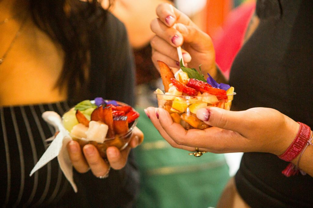 다채로운 과일 샐러드와 딸기, 자두, 식용 꽃을 담은 그릇을 들고 있는 두 사람, 한 사람은 숟가락으로 음식을 먹습니다.