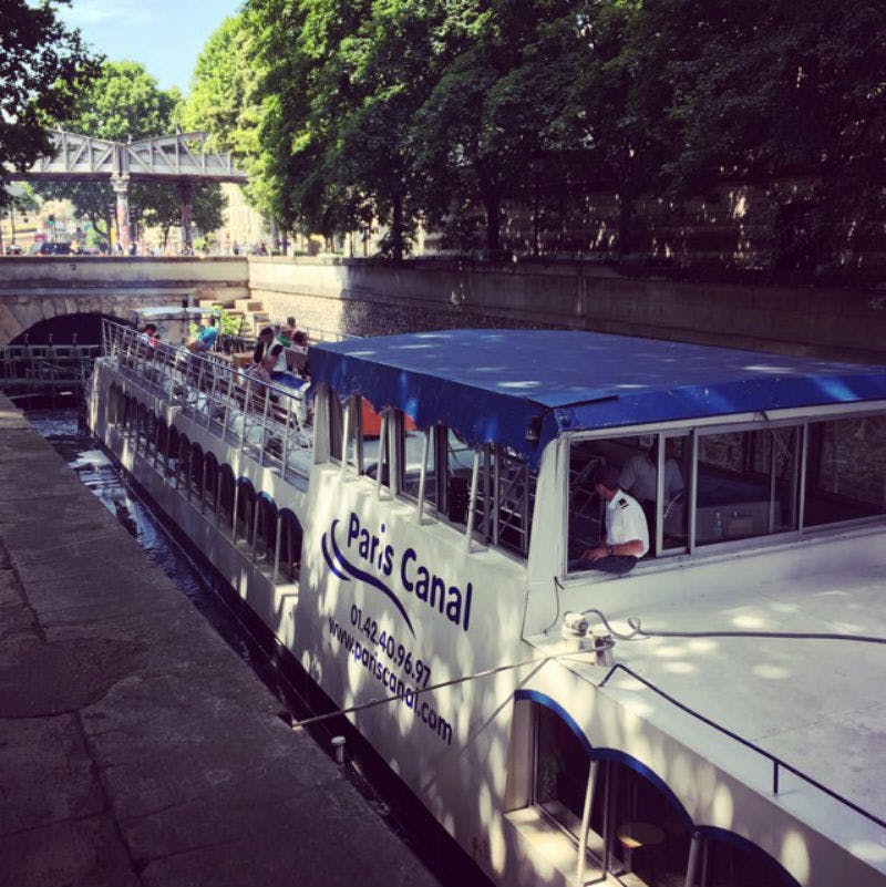 "파리 운하" 라는 이름의 관광 보트가 다리 아래 나무와 돌담으로 둘러싸인 운하를 항해합니다.