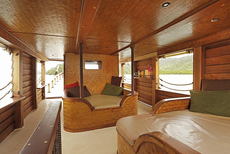 목재 및 대나무 가구, 쿠션이 있는 좌석, 물과 언덕의 경치를 감상할 수 있는 창문이 있는 하우스보트 내부.