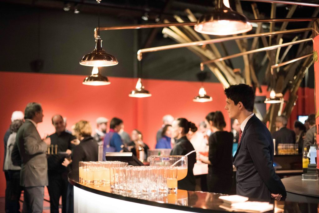 정장을 입은 바텐더가 음료수와 유리잔을 들고 바 뒤에 서 있으며, 따뜻한 불이 켜진 장소에서 사람들과 어울리고 있습니다.