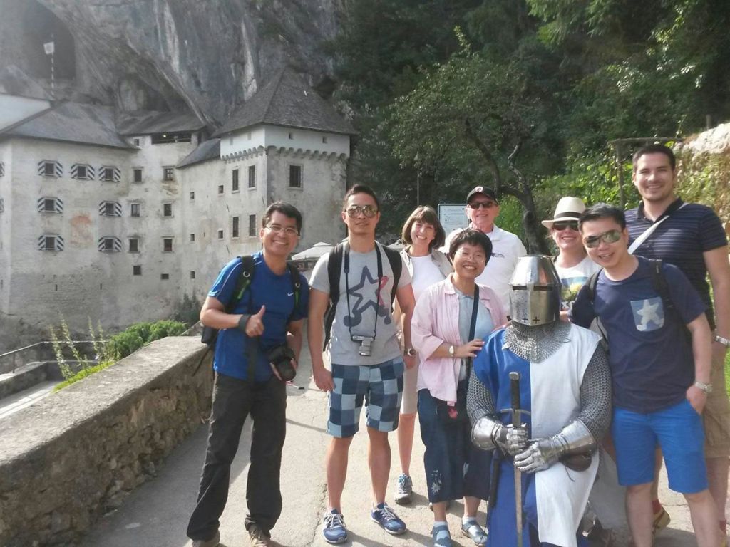 한 무리의 사람들이 소박한 건물과 녹지 앞에서 기사의 갑옷을 입은 사람과 포즈를 취하고 있다.