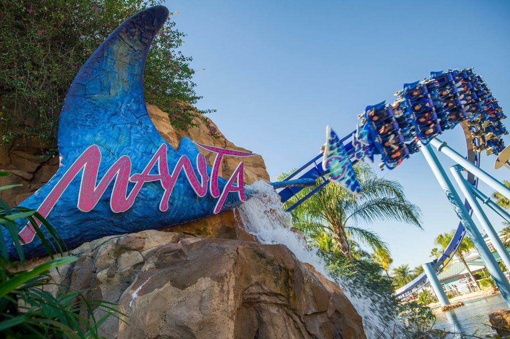 '만타' 표지판이 있는 만타 레이를 테마로 한 롤러코스터, 바위 위로 흐르는 물, 파란색 트랙을 배경으로 달리는 사람들.