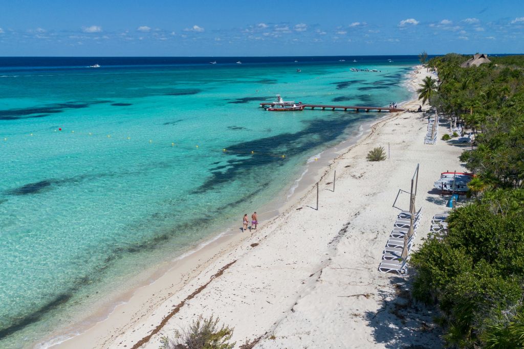 희박한 초목, 맑고 푸른 물, 부두, 몇 척의 보트, 해변을 걷는 두 사람이 있는 모래 해변의 공중 전망.