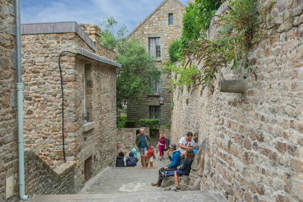 녹음이 우거진 소박한 석조 건물 사이 좁은 돌길을 걸으며 앉아 있는 사람들.