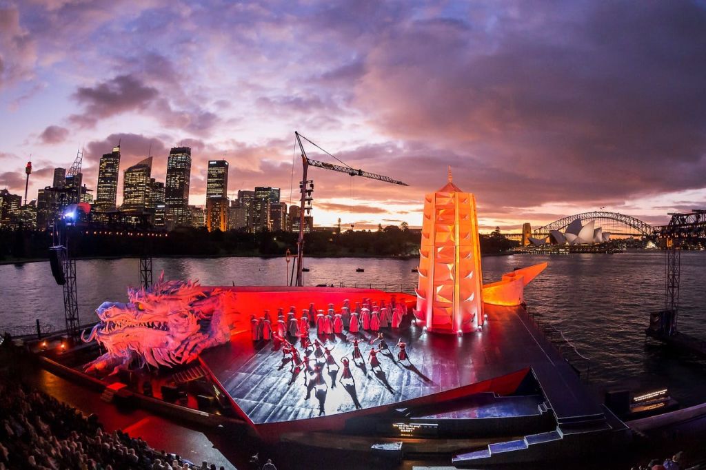 무용수, 붉은 탑, 도시의 스카이라인, 석양이 비치는 하늘을 배경으로 펼쳐지는 해안가 무대에서 공연을 즐겨보세요.