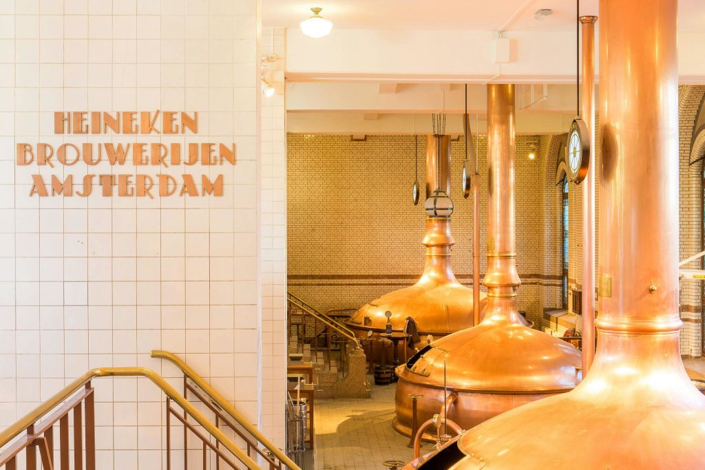 왼쪽 벽에 구리 양조 탱크, 흰색 타일 벽, "Heineken Brouwerijen Amsterdam" 텍스트가 있는 양조장 내부.