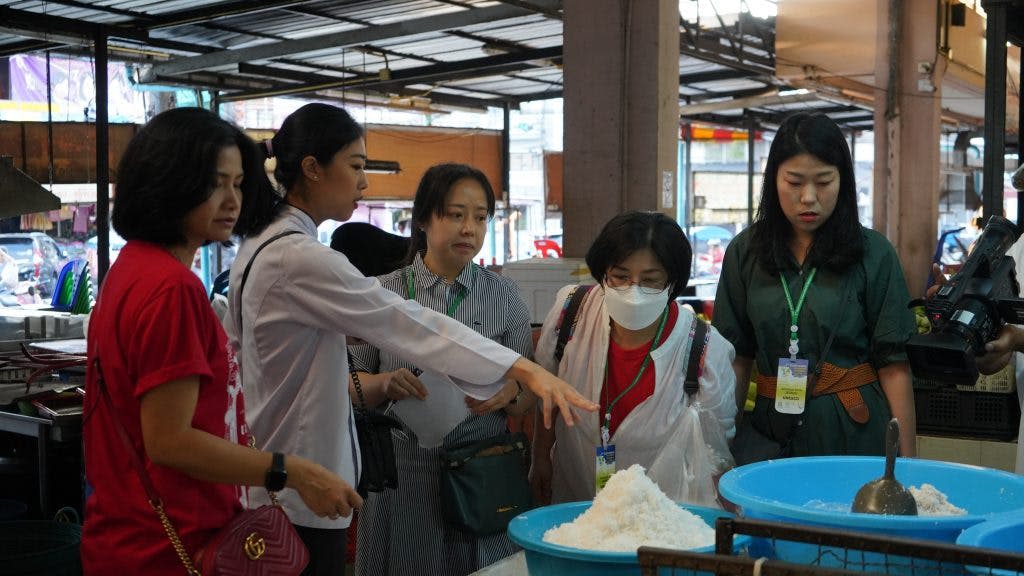 세 사람이 실내 시장에서 파란색 용기에 하얀 물질이 쌓여 있는 것을 관찰하고, 한 사람은 가리키고 다른 한 사람은 필름을 찍습니다.