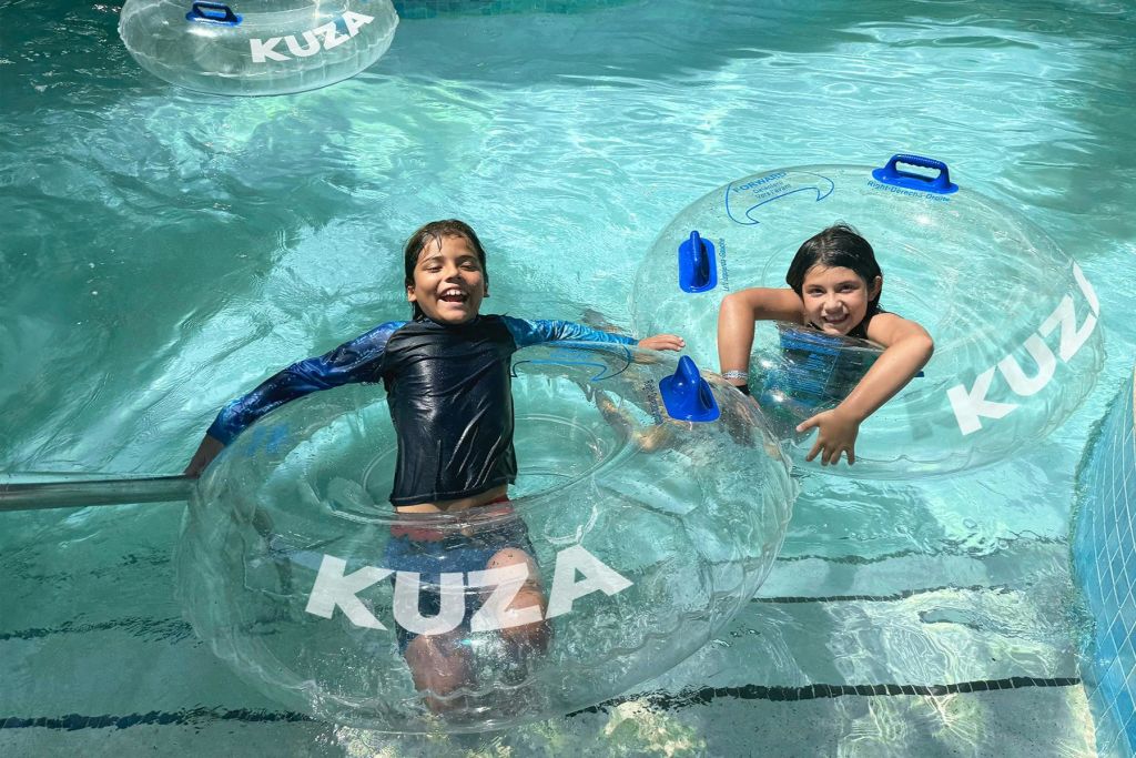 "KUZA" 라고 표시된 투명한 팽창식 튜브가 있는 야외 수영장에서 두 어린이가 미소 짓고 놀고 있습니다.