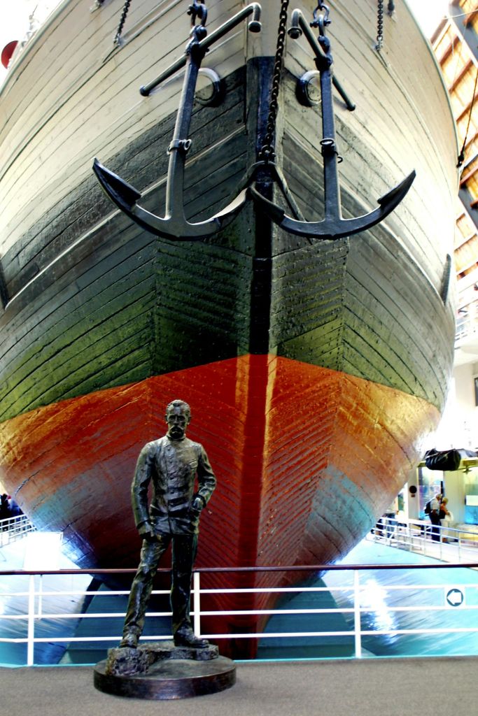 난간 위에 실내에 전시된 듀얼 앵커가 있는 대형 선박 활. 해군 복장을 한 남자의 동상이 정면에 서 있습니다.