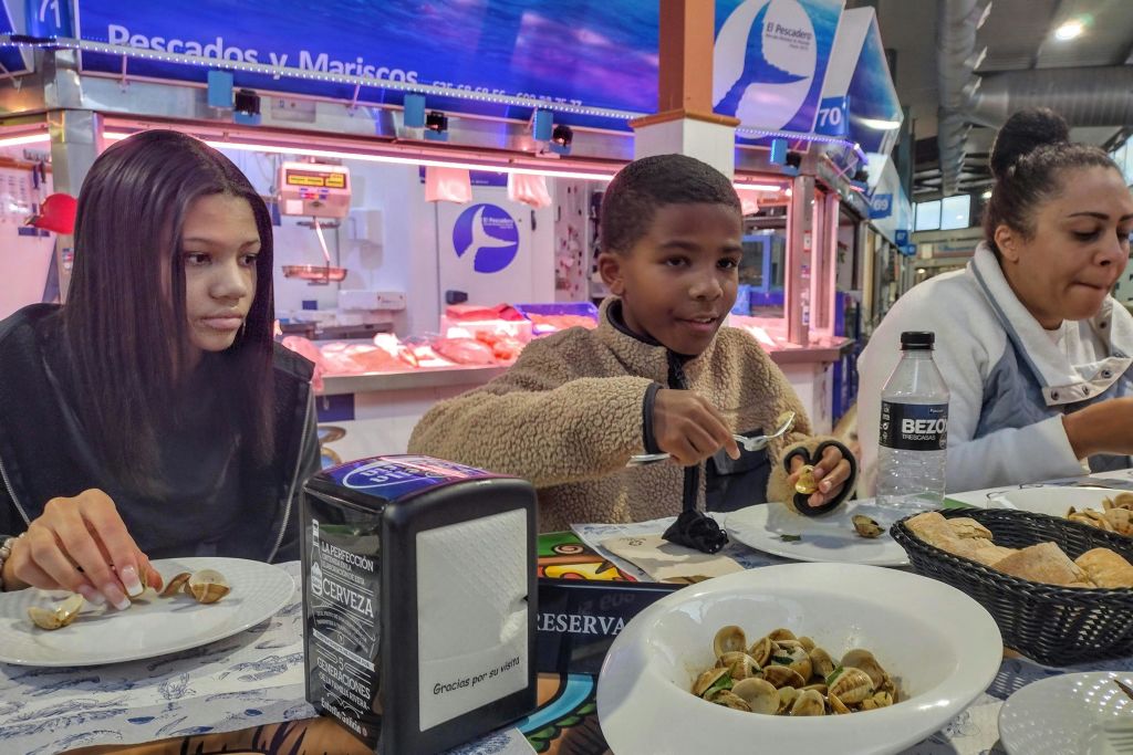 두 어린이가 레스토랑 테이블에서 "Pescados y Mariscos" 라고 적힌 표지판을 배경으로 해산물을 먹고 있습니다.