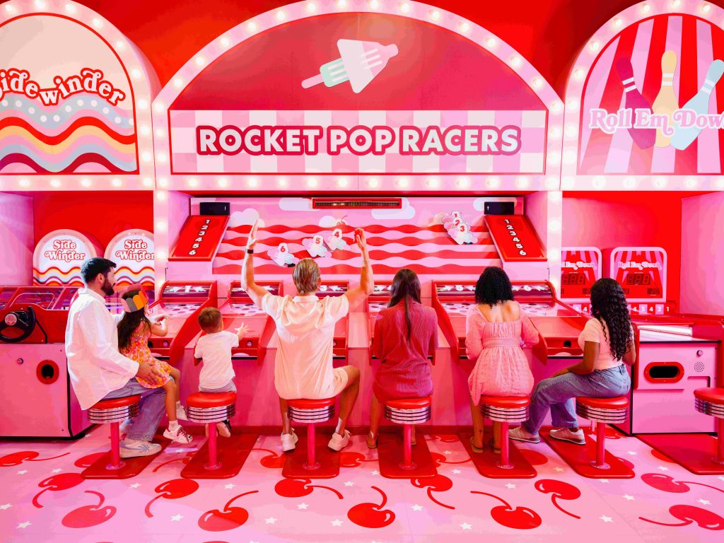 밝은 색상의 체리 테마 룸에서 열리는 로켓 팝 레이서 (Rocket Pop Racers) 아케이드 게임에서 두 명의 어린이를 포함한 6명이 빨간 의자에 앉아 있습니다.