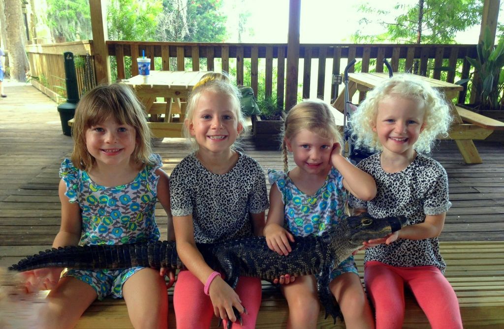 표범 프린트 상의와 분홍색 바지를 입은 세 명의 어린 소녀가 피크닉 테이블이 있는 나무 데크에 앉아 작은 악어를 들고 있습니다.