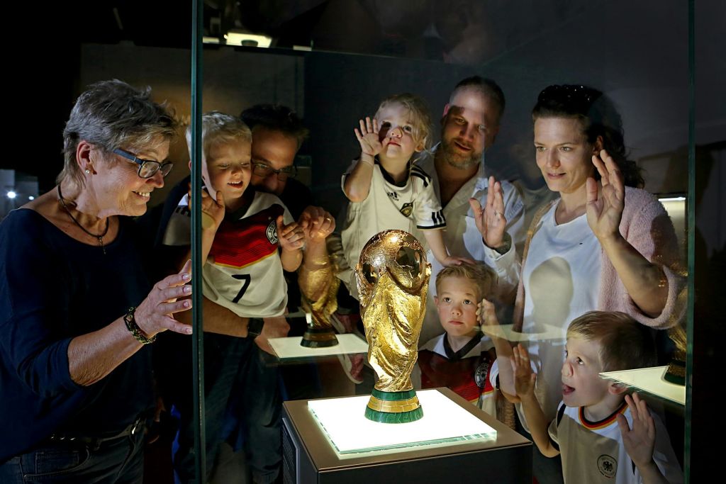 축구 유니폼을 입은 어린이를 포함한 몇몇 사람들은 유리 디스플레이 케이스 안에 불이 켜진 월드컵 트로피를 봅니다.