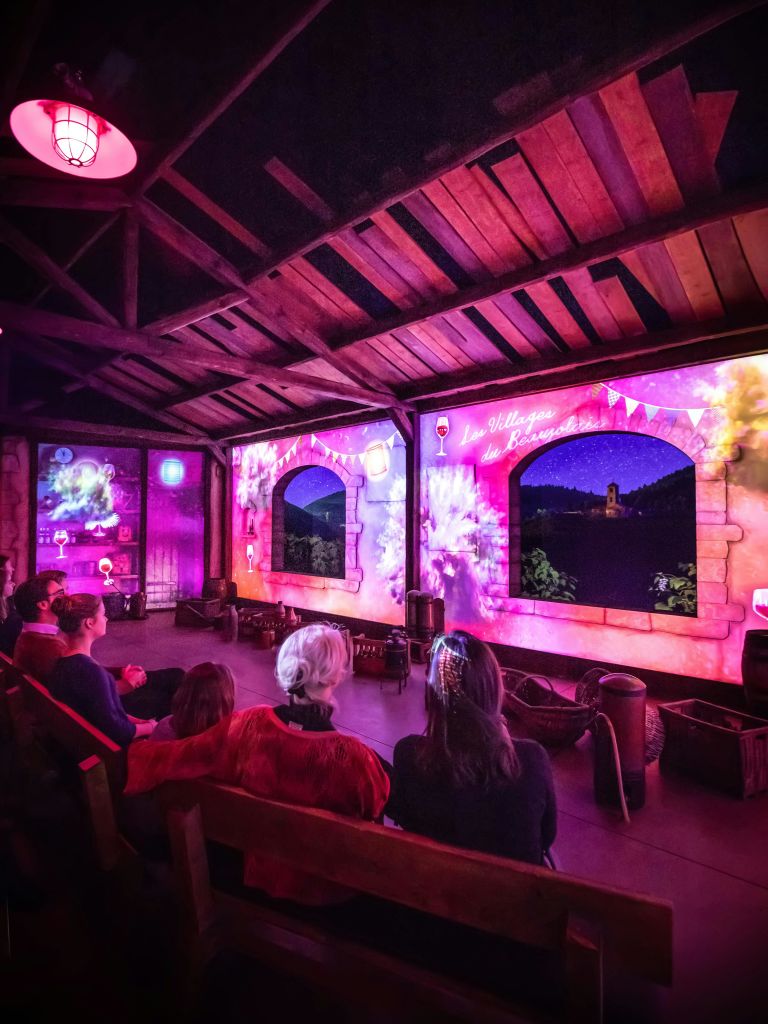 조명이 희미한 방에 앉아 나무 기둥 천장 아래에서 창문을 닮은 벽의 다채로운 투사 장면을 보고 있는 사람들.