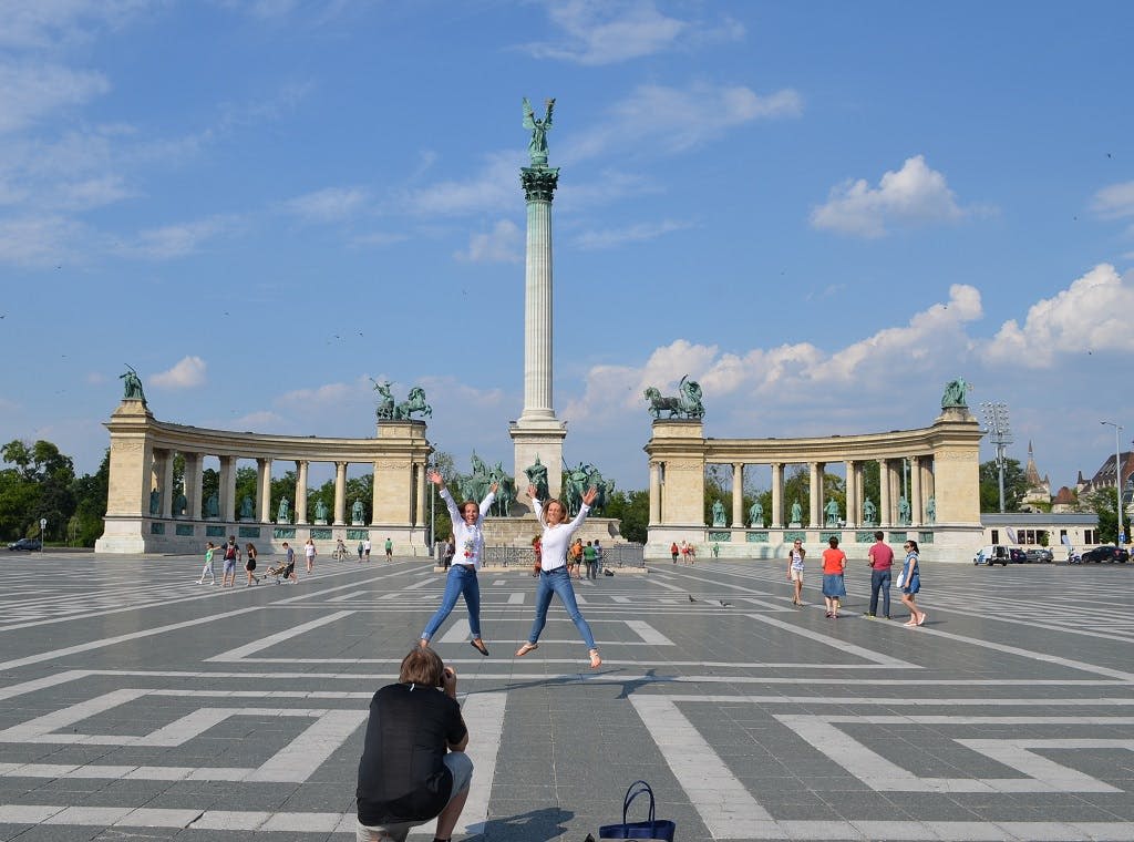 석상이 있는 커다란 기념비 앞에서 사진을 찍기 위해 포즈를 취하는 동안 두 사람이 공중으로 뛰어오르고, 다른 사람들은 뒤에서 돌아다닙니다.