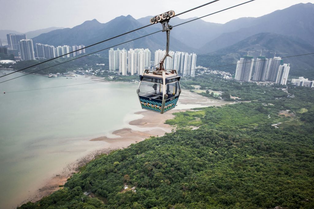 높은 건물과 산을 배경으로 푸른 숲과 만 위로 케이블카가 운행됩니다.