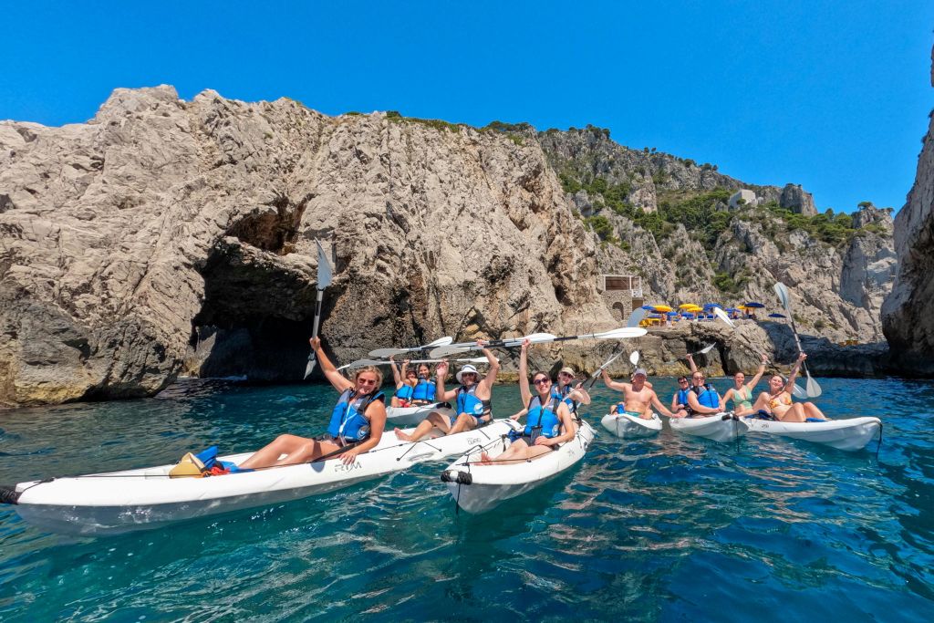 밝고 푸른 하늘 아래 바위 절벽 근처의 맑고 푸른 물에서 구명조끼를 입고 카약을 타는 사람들.