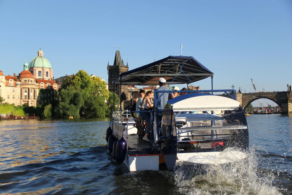 맑고 푸른 하늘 아래 역사적인 건물과 높은 탑이 보이는 강에서 캐노피로 덮인 보트를 타는 사람들이 있습니다.