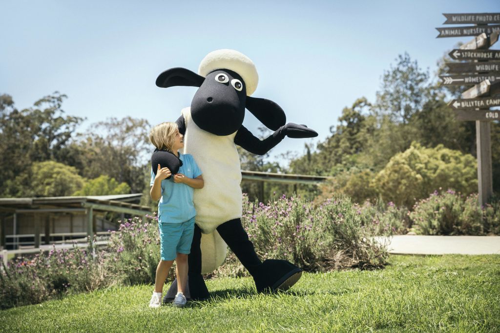 나무와 덤불을 배경으로 한 야외 공원에서 푸른 옷을 입은 어린이가 양 의상을 입은 큰 사람을 껴안고 있습니다.