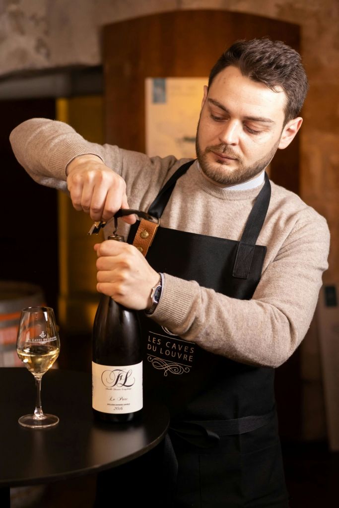 스웨터를 입고 앞치마를 두른 남성이 코르크 나사가 달린 와인병을 따듯한 조명으로 열고 있습니다.