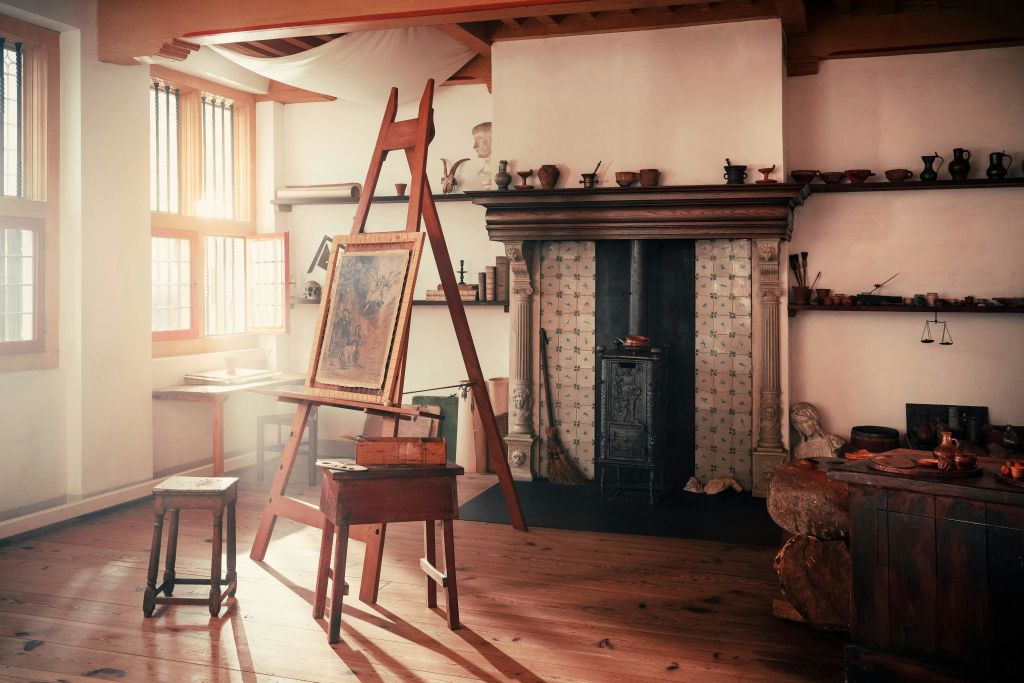 이젤, 페인팅, 목재 스툴, 선반, 벽난로가 있는 소박한 아트 스튜디오입니다. 햇빛이 창문을 통해 흐릅니다.