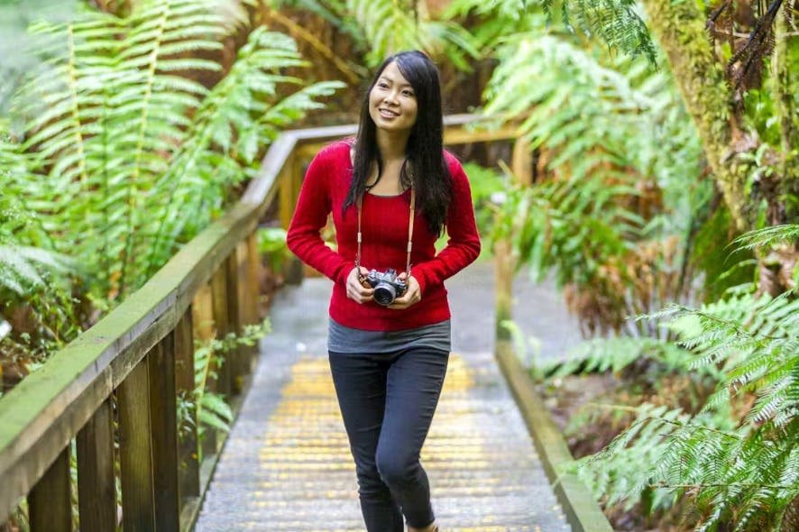 붉은 스웨터를 입은 여성이 숲 속 푸른 양치류로 둘러싸인 나무 통로를 걷는 동안 카메라를 들고 있습니다.