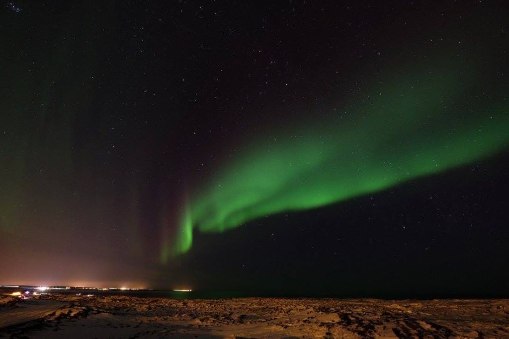 수평선에 조명이 보이는 눈 덮인 풍경 위로 별이 빛나는 밤하늘을 가로지르는 녹색 오로라 보레알리스.