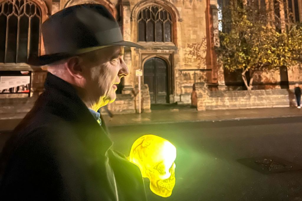 어두운 코트와 모자를 쓴 사람이 밤에 역사적인 건물 근처에서 빛나는 노란색 두개골을 들고 있습니다.