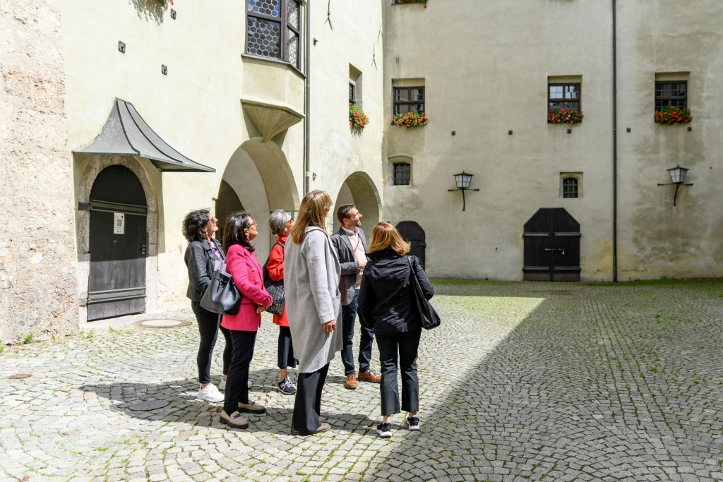 아치형 출입구와 꽃 상자가 있는 유서 깊은 건물로 둘러싸인 자갈 마당에 한 무리의 사람들이 서 있습니다.