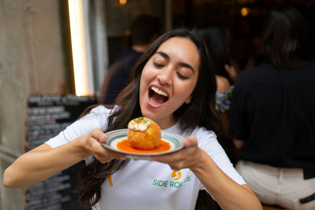 "사이드 로드" 로고가 있는 흰색 셔츠를 입고 빨간색 소스 침대에 튀긴 공과 함께 요리를 먹으려는 미소를 짓는 여성.