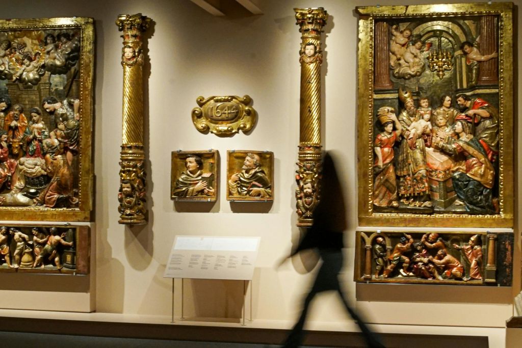 박물관에서 금색 액자로 장식된 화려한 종교 예술품과 조각품이 전시된 벽면 너머로 흐릿한 인물이 걸어가고 있습니다.