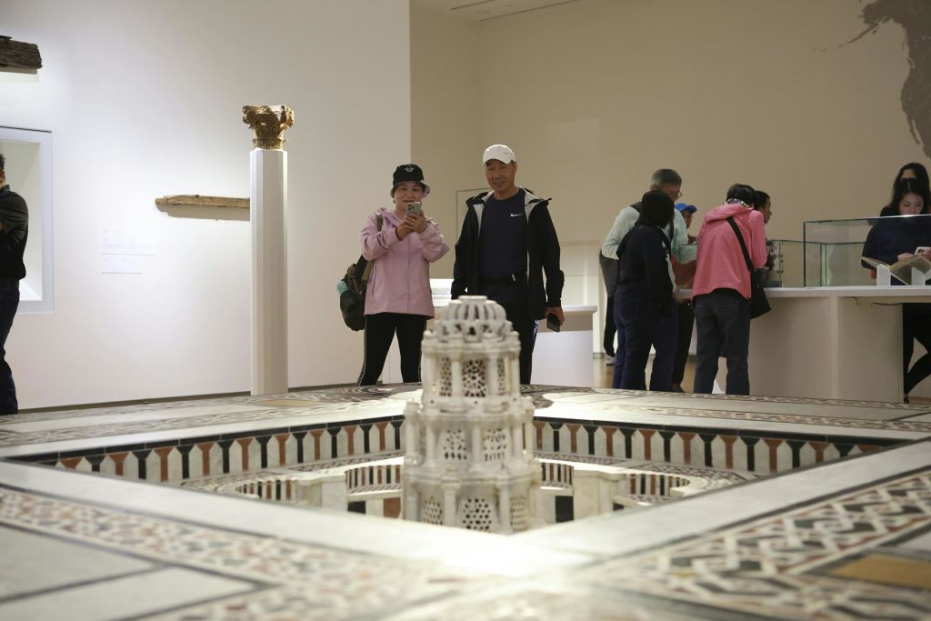 사람들은 박물관 전시관에서 미니어처 건축 모델을 관찰합니다. 다른 유물은 배경의 받침대에 표시됩니다.
