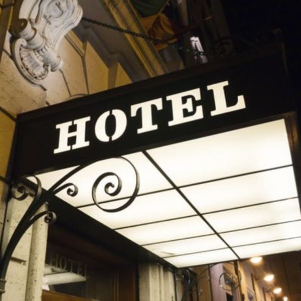 입구 위에 장착된 화려한 금속 브래킷이 있는 조명 호텔 표지판. 건물 세부 정보가 배경에 표시됩니다.