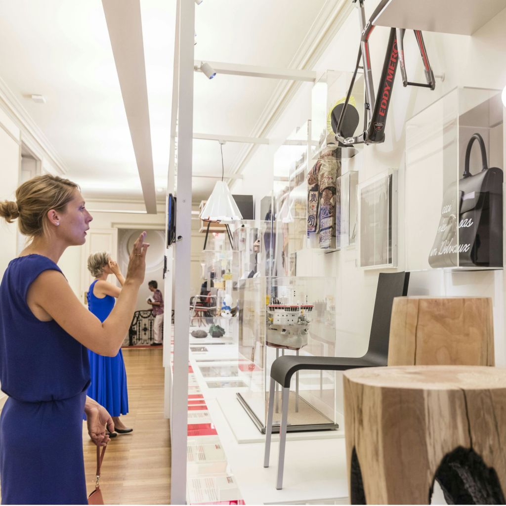 파란색 드레스를 입은 여성이 현대적인 갤러리의 디스플레이와 상호 작용하며, 선반에 다양한 물건이 있고 벽에 자전거가 장착되어 있습니다.