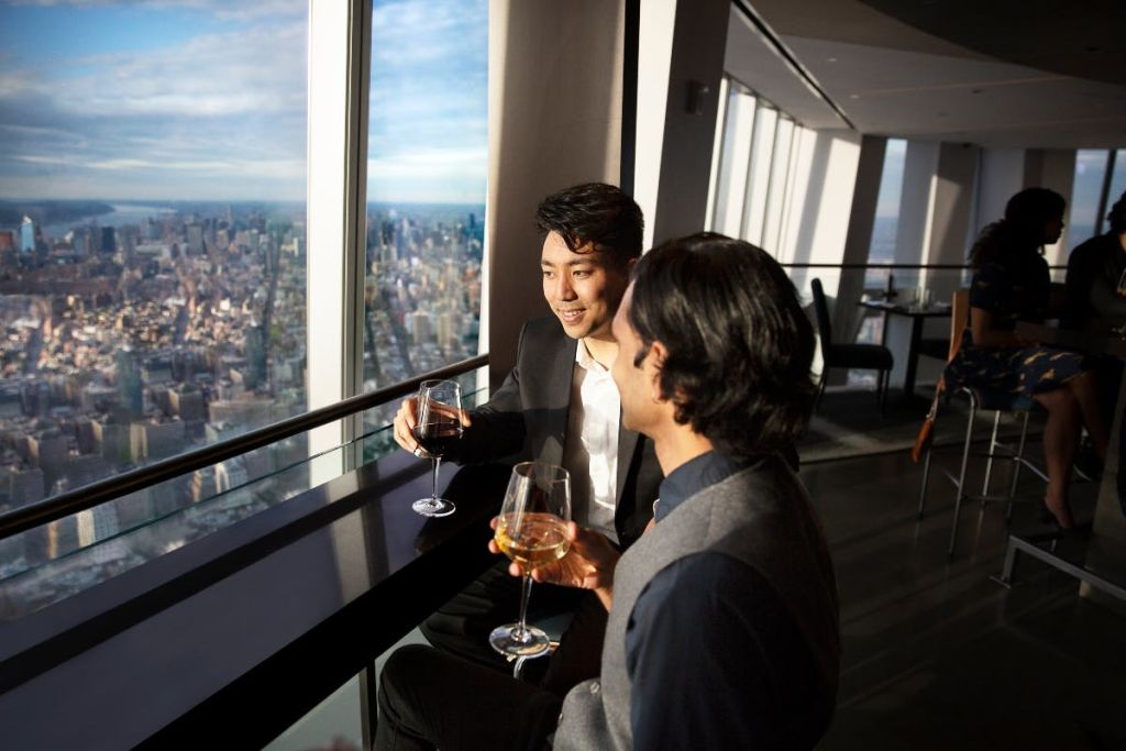 고층 레스토랑의 두 사람이 커다란 창문을 통해 도시 풍경을 감상하며 와인잔을 즐기고 있습니다.