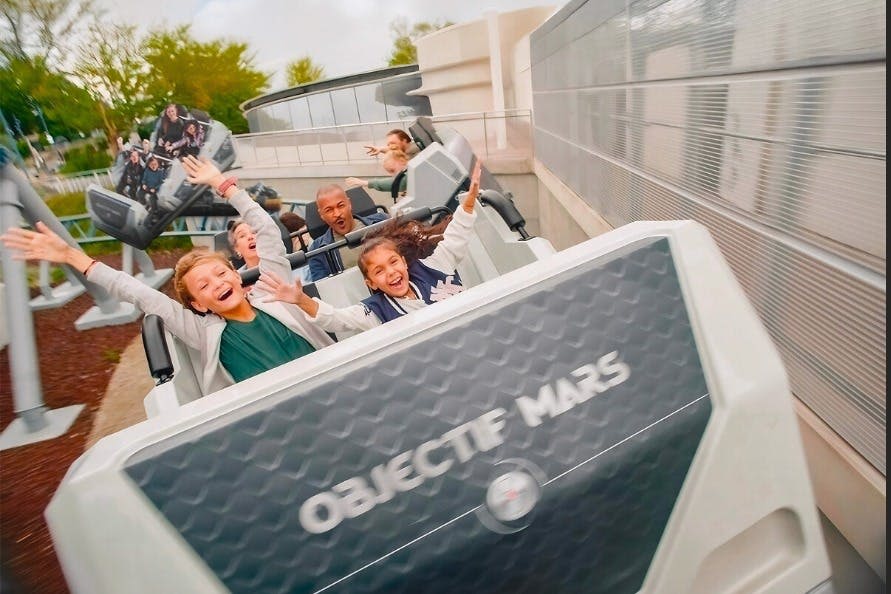 나무와 현대적인 구조로 둘러싸인 두 팔을 들고 롤러코스터를 타는 사람들을 만나보세요.