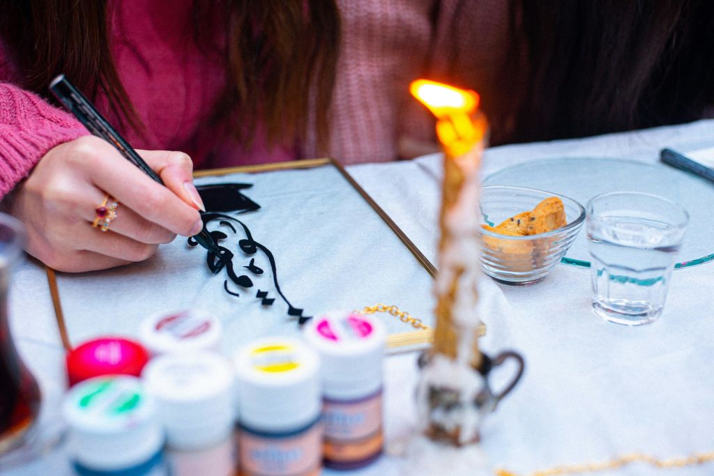 화사한 페인트, 불타는 촛불, 근처에 쿠키 한 그릇이 있는 테이블에 검은색 페인트로 유리 위에 그림을 그리는 사람.