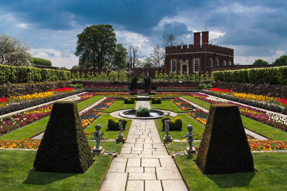중앙 분수대, 대칭형 화단, 울타리, 동상, 흐린 하늘 아래 벽돌 건물이 배경으로 있는 정식 정원입니다.