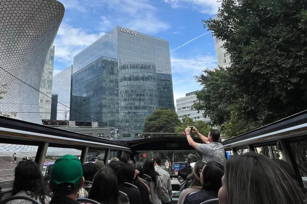 "Telcel" 이라는 라벨이 붙은 건물과 푸른 하늘 아래 나무를 포함한 유리 건물이 있는 도시에서 노천 버스 투어를 하는 사람들.