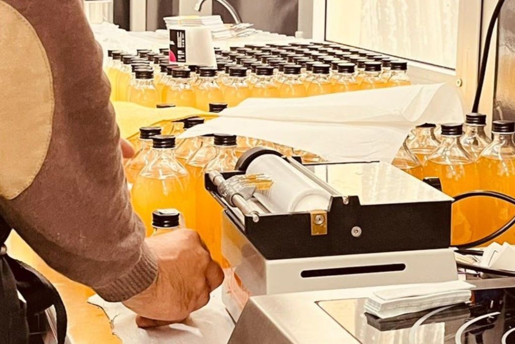 한 사람이 수많은 주황색 액체 병이 채워진 테이블에서 기계를 작동합니다. 각 병에는 검은색 뚜껑이 있습니다.