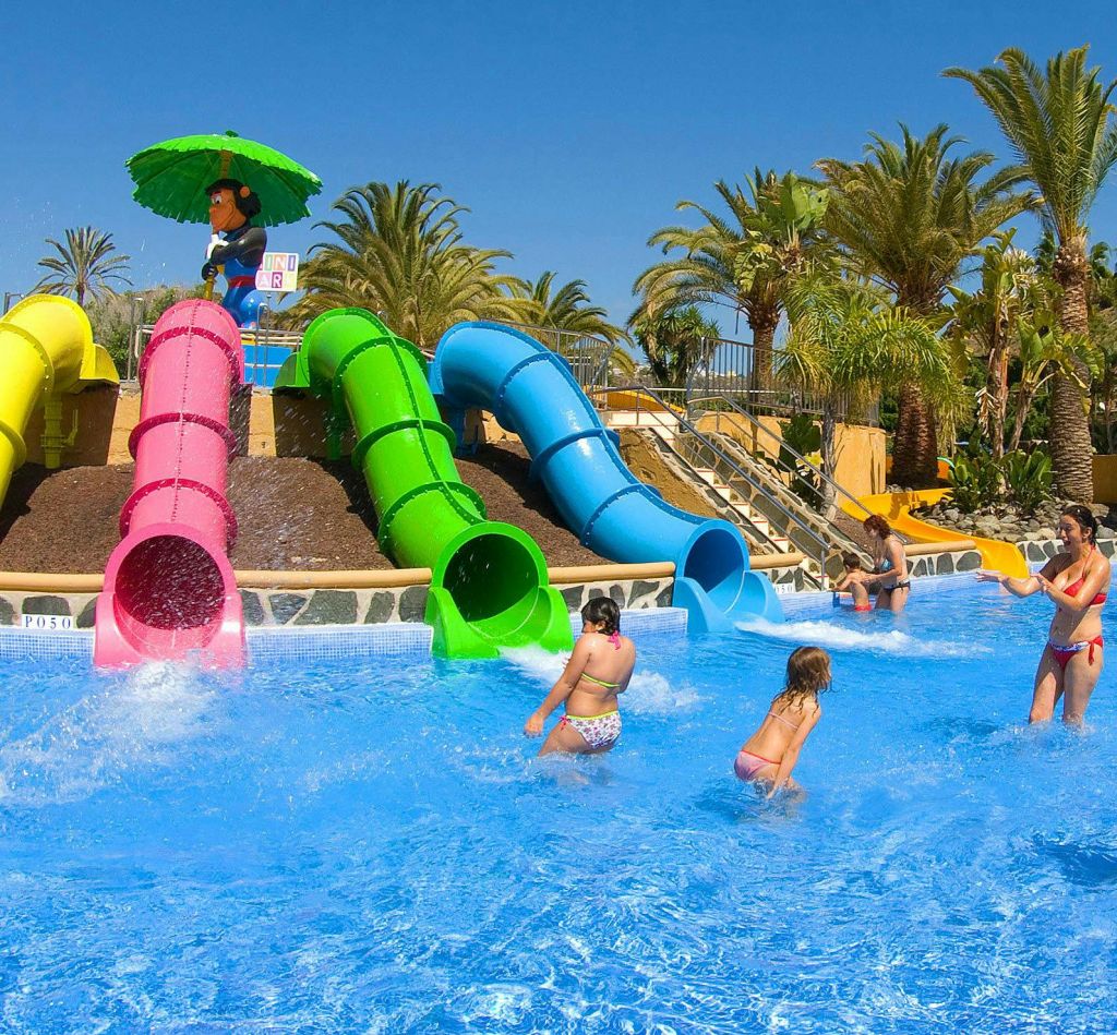 화창한 날 야자수로 둘러싸인 세 개의 다채로운 워터 슬라이드 (분홍색, 녹색, 파란색) 가 있는 수영장에서 놀고 있는 어린이들.