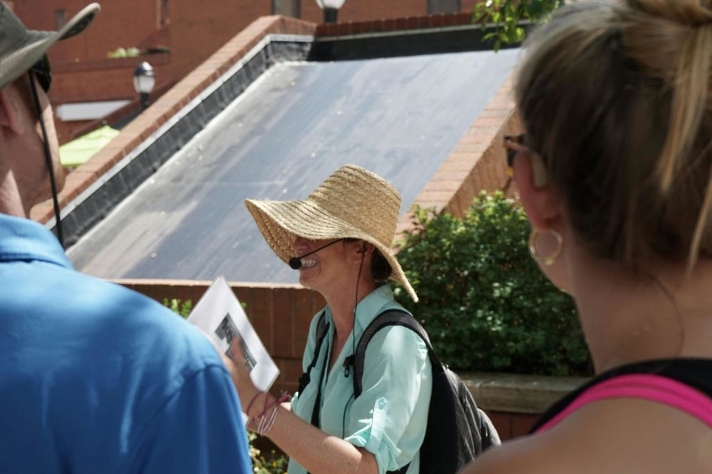 밀짚모자와 마이크 헤드셋을 착용하고 종이를 들고 벽돌 건물과 녹지 근처에서 야외에서 말하는 사람.