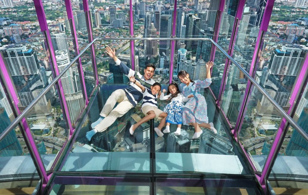 4인 가족이 고층 전망대의 유리 바닥에 누워 있으며, 아래와 주변에 도시 풍경이 보입니다.