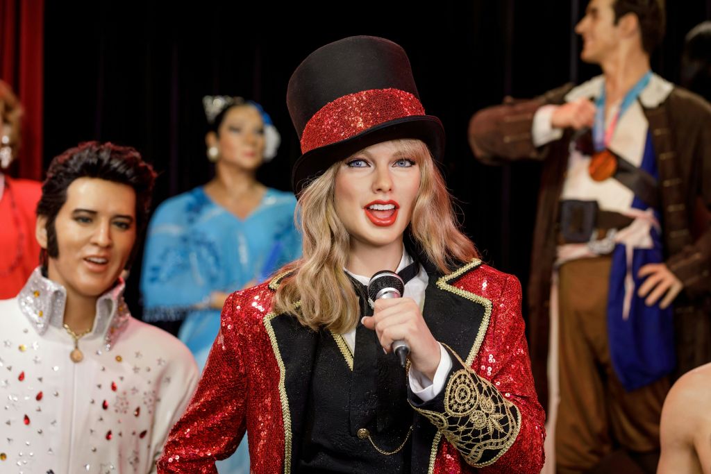 빨간색 스팽글 재킷과 상의를 입은 사람이 마이크를 들고 있으며, 의상을 입은 흐릿한 형상이 배경에 있습니다.