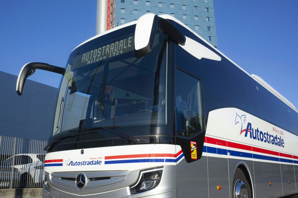 디지털 표지판에 "Milano 1924" 가 표시된 현대적인 Autostradale 버스는 높고 패턴이 있는 건물 앞에 주차되어 있습니다.