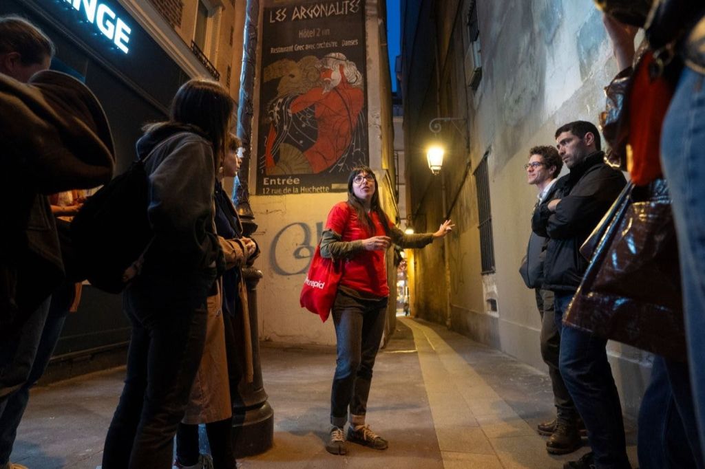 빨간색 셔츠를 입은 여성이 좁은 골목에서 한 무리의 사람들과 이야기를 나누고 있으며, 빈티지 가로등이 현장을 비추고 있습니다.
